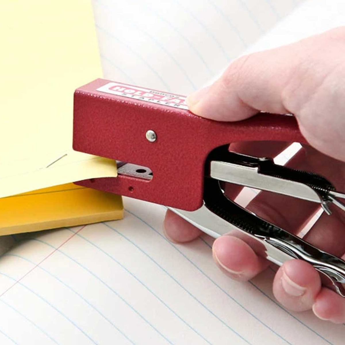 Red stapler being used on a piece of paper with a notebook in the background
