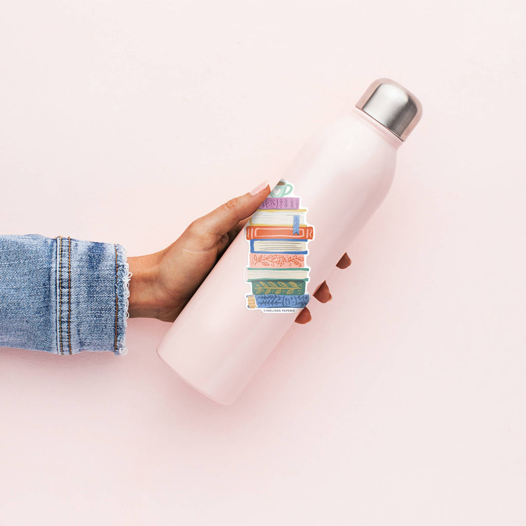 Person holding a pink water bottle with colorful vinyl sticker of a bookstack on a light pink background