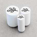 Three white cylindrical stamps with leaf designs on a gray background