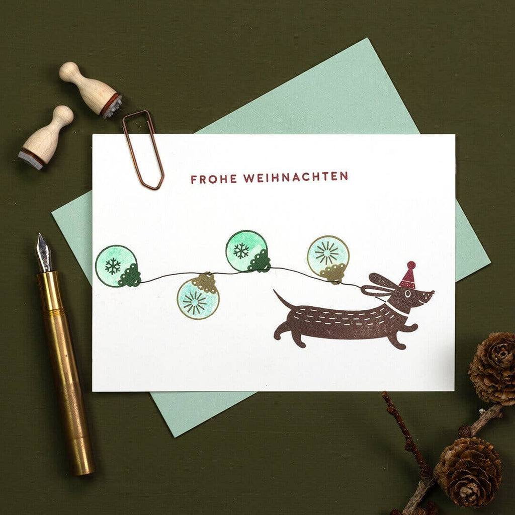 Christmas card with a dachshund and lights, surrounded by stationery items on a dark surface.