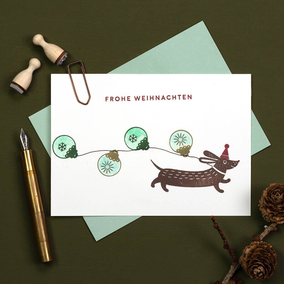 Christmas card with a dachshund and lights, surrounded by stationery items on a dark surface.