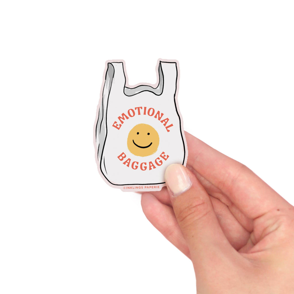 Vinyl sticker of Small white bag with 'Emotional Baggage' text and smiley face held by a hand on a white background