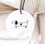 Round white ornament with a dachshund illustration and heart, hanging against a textured beige background.