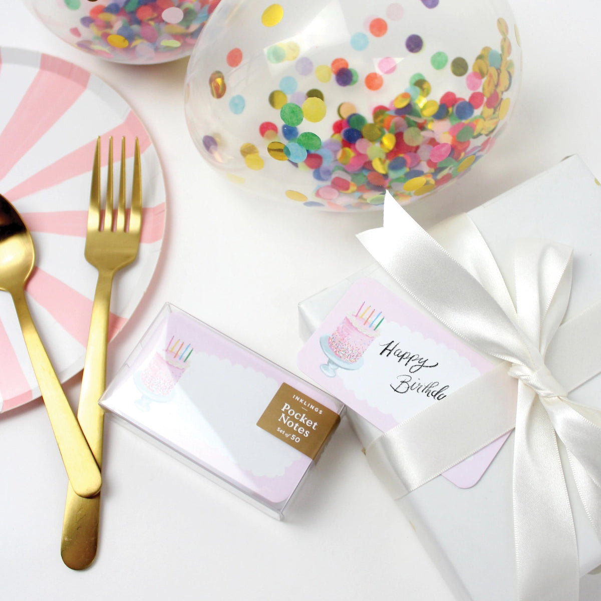 Table setting with birthday-themed items including a box of pocket notes, a fork, knife, and colorful confetti plates.