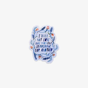A die-cut sticker featuring the hand-lettered quote 'I WILL NOT LOSE THIS MOMENT SEARCHING FOR ANOTHER' in dark blue, surrounded by a watercolor-style floral wreath in shades of blue, purple, and peach, on a light gray background.