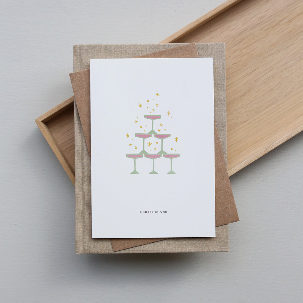 Greeting card with a cake illustration and 'A toast to you' text, placed on a brown envelope with a wooden tray in the background.