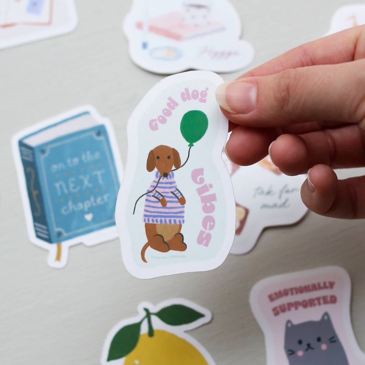 Hand holding a vinyl sticker with a dog, balloon, and the text 'good dog vibes',  surrounded by other colorful stickers on a light surface.