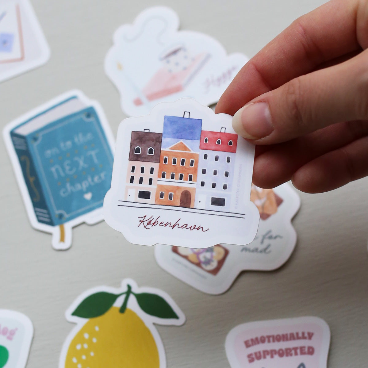 Hand holding a sticker with a Nyhavn cityscape design and 'København' text, surrounded by other colorful stickers on a light surface.