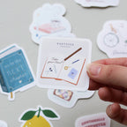 Hand holding a vinyl sticker with a journal filled with journaling illustrations, a pencil writing 'take your time' and a Kartotek Copenhagen logo, on a surface with other stickers.