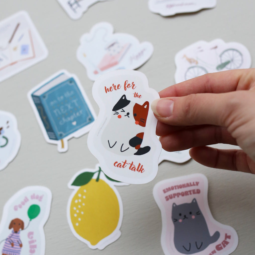 Hand holding a sticker with cat illustrations and text, surrounded by other colorful stickers on a light background