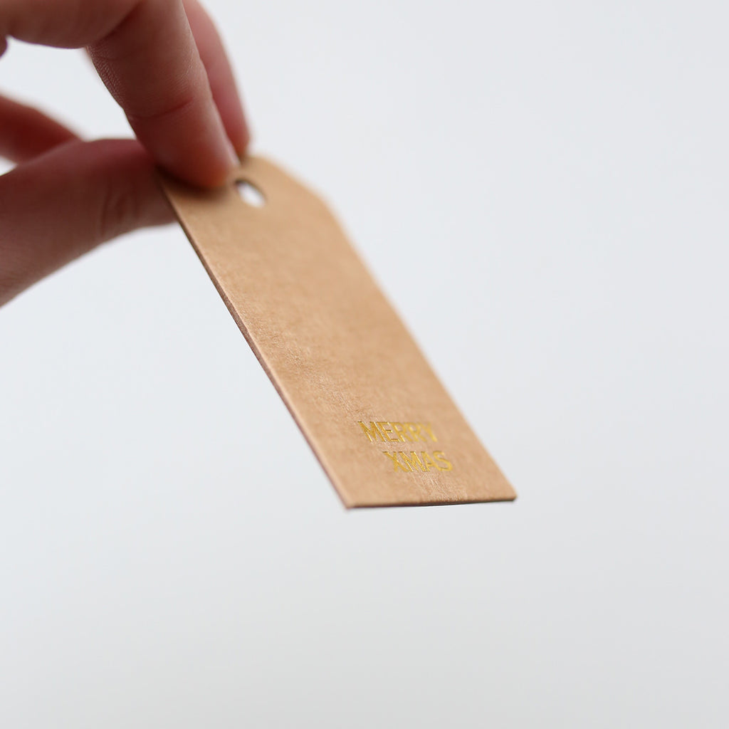 Hand holding a brown gift tag with gold 'Merry xmas' text on a white background
