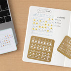 Notebook with calendar stencils on a wooden surface next to a laptop.