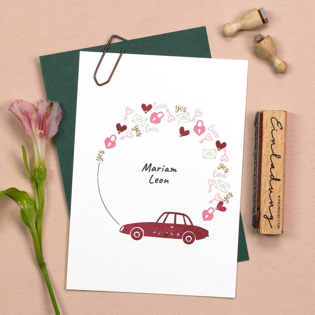 Greeting card with car and heart designs on a beige surface with flowers and stamps.
